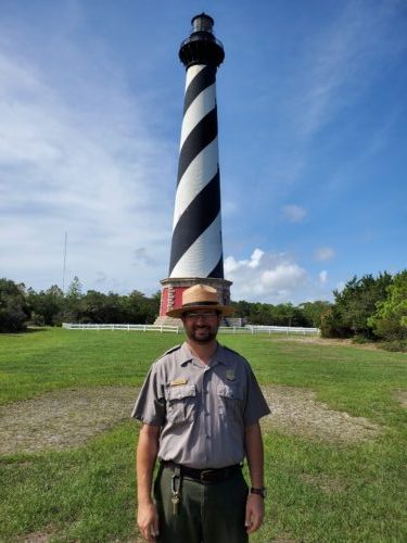 Ranger Jonathan stands in front of the Cape Hatteras Lighthouse, which has white and black stripes and a red base.
