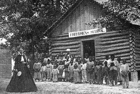 A historical black and white photograph of Freedmen's Colony students and their teacher standing in front of the Freedmen's School on Roanoke Island.