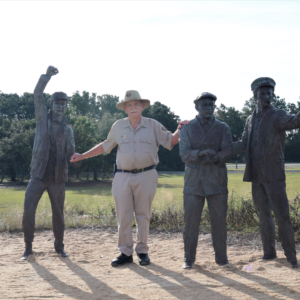 Volunteer Fred Hattman stands with Wright Brothers statues at Wright Brothers National Memorial on a sunny day 