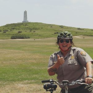 A female park ranger gives the thumbs up sign while sitting on a bicycle with Wright Brothers National Memorial in the background. 