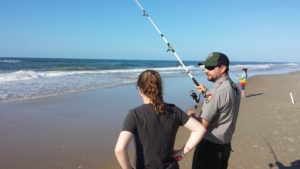 A male ranger holds a fishing pole while talking to a female visitor on the beach at Cape Hatteras National Seashore with the ocean in the background.
