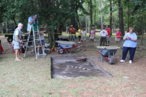 A group of archaeologists excavates a rectangular section of earth on the grounds of Fort Raleigh National Historic Site while visitors watch.