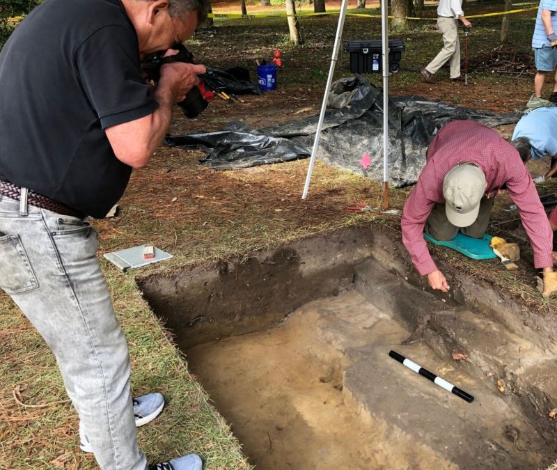 An archaeologist examines a rectangular section of earth at Fort Raleigh National Historic Site while a photographer takes a picture.