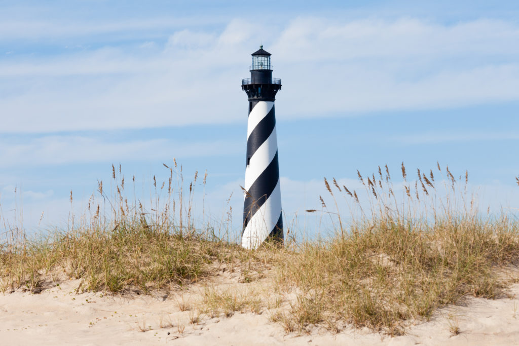 The Cape Hatteras Lighthouse photographed against a blue sky with clouds on a sunny day with a sand dune and sea oats in the foreground.