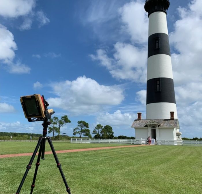 A vintage camera on a tripod in front of the black and white striped Bodie Island Lighthouse.