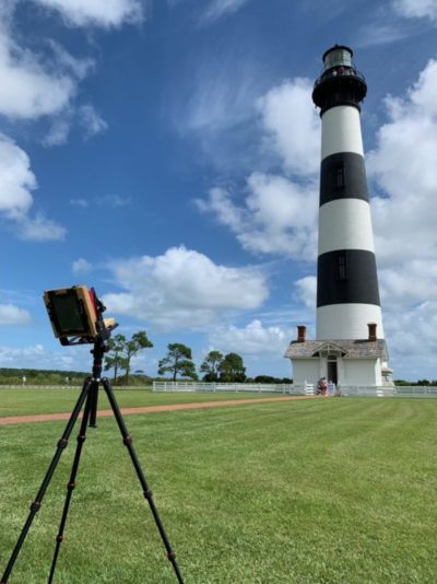 A vintage camera on a tripod in front of the black and white striped Bodie Island Lighthouse.