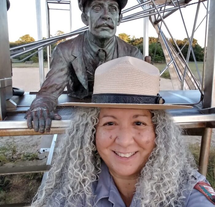 Ranger Lulu Luis in front of a sculpture of Orville Wright on the 1903 Wright Flyer at Wright Brothers National Memorial.