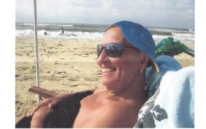 Barbara Jordan, wearing a sideways baseball hat and sunglasses and smiling, seated in a beach chair in the sand on Cape Hatteras National Seashore. An umbrella pole and a person playing in the ocean can be seen in the background.