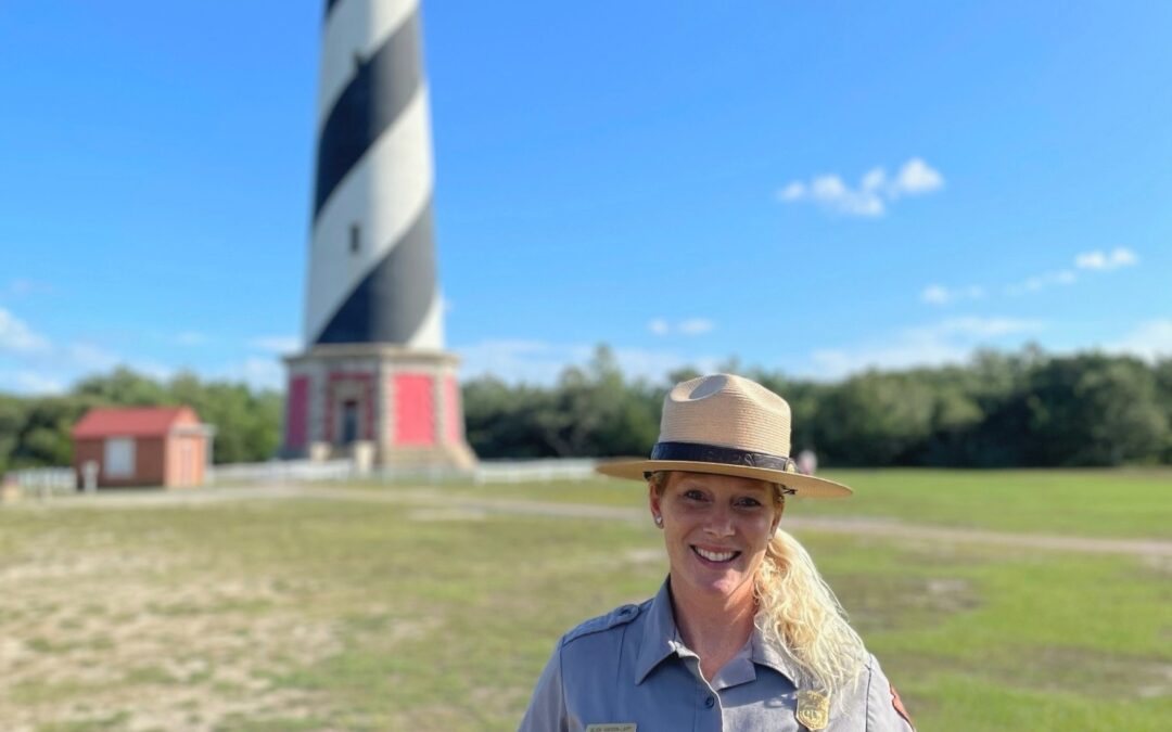 Ranger Alisa Hardin-Lapp on the grounds of the Cape Hatteras Lighthouse. The black-and-white diagonally striped lighthouse and an outbuilding can be seen in the background.