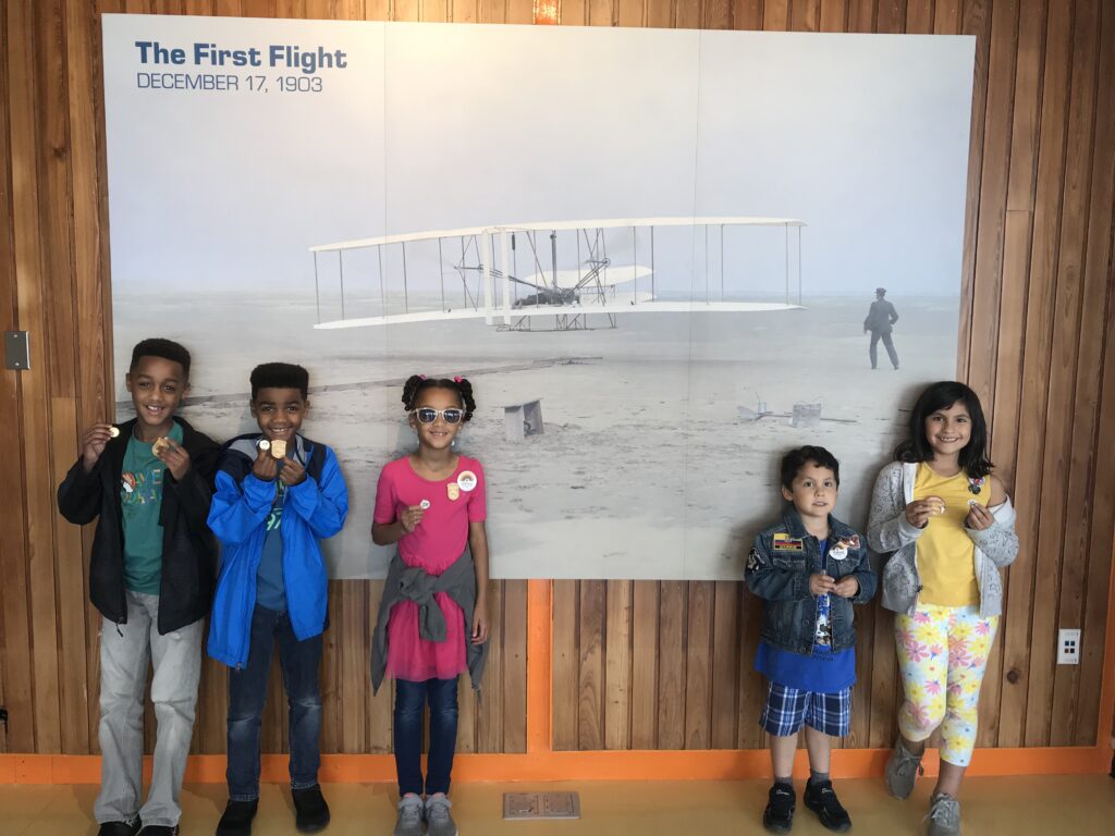 Five Junior Rangers (three on left, two on right) stand in front of the first flight photograph at Wright Brothers National Memorial.