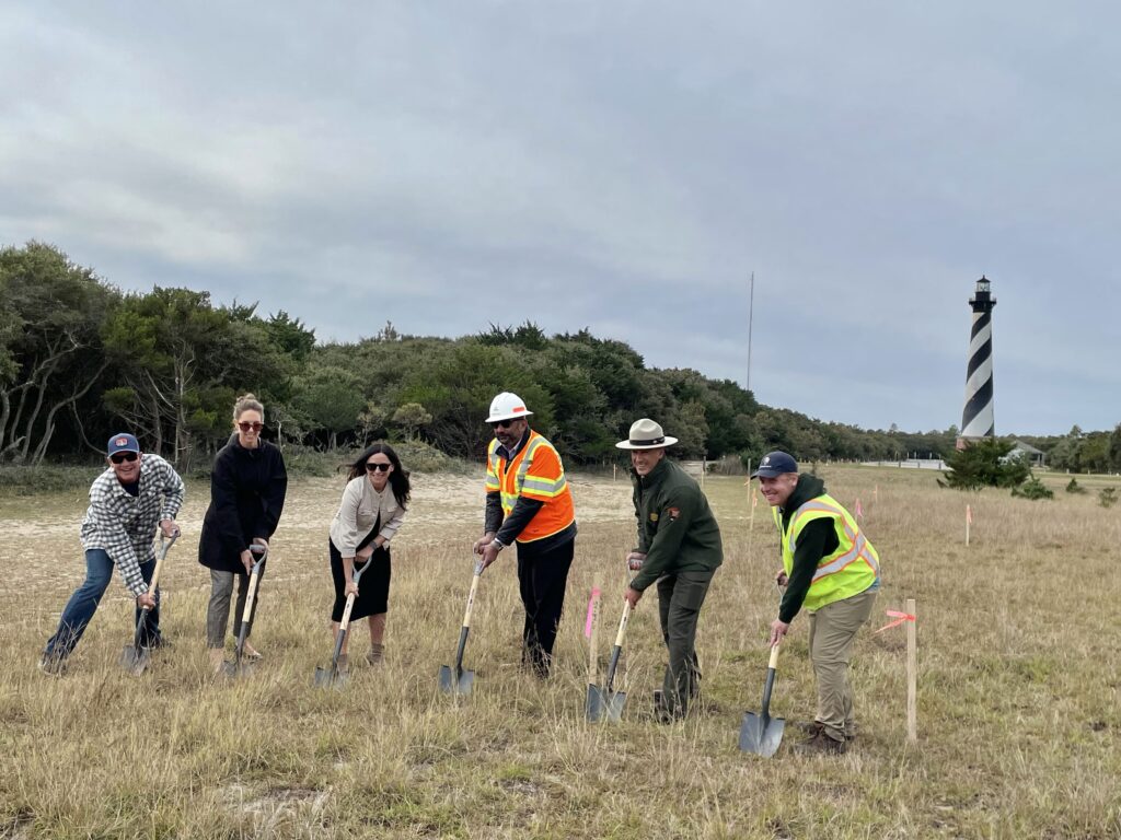 A photograph of six people holding shovels pointed into the ground. Pictured from left to right: Trip Forman, Outer Banks Forever Board President and Co-Owner of REAL Watersports; Laura Ertle, Director of Marketing and Public Relations at Cape Hatteras Electric Cooperative; Jessica Barnes, Director of Outer Banks Forever; Babur Mian, President and CEO of Terra Site Constructors, LLC; David Hallac, Superintendent of the National Parks of Eastern North Carolina; and Steven Torgerson, Landscape Architect with the National Park Service. The Cape Hatteras Lighthouse can be seen in the background.