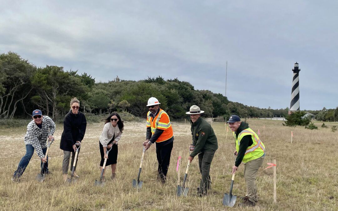A photograph of six people holding shovels pointed into the ground. Pictured from left to right: Trip Forman, Outer Banks Forever Board President and Co-Owner of REAL Watersports; Laura Ertle, Director of Marketing and Public Relations at Cape Hatteras Electric Cooperative; Jessica Barnes, Director of Outer Banks Forever; Babur Mian, President and CEO of Terra Site Constructors, LLC; David Hallac, Superintendent of the National Parks of Eastern North Carolina; and Steven Torgerson, Landscape Architect with the National Park Service. The Cape Hatteras Lighthouse can be seen in the background.