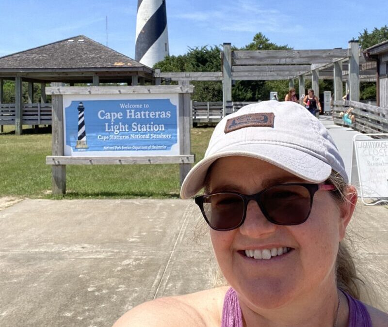 Shannon Winakur takes a selfie in front of the sign for the Cape Hatteras Light Station. The black and white diagonally striped Cape Hatteras Lighthouse can be seen in the background.
