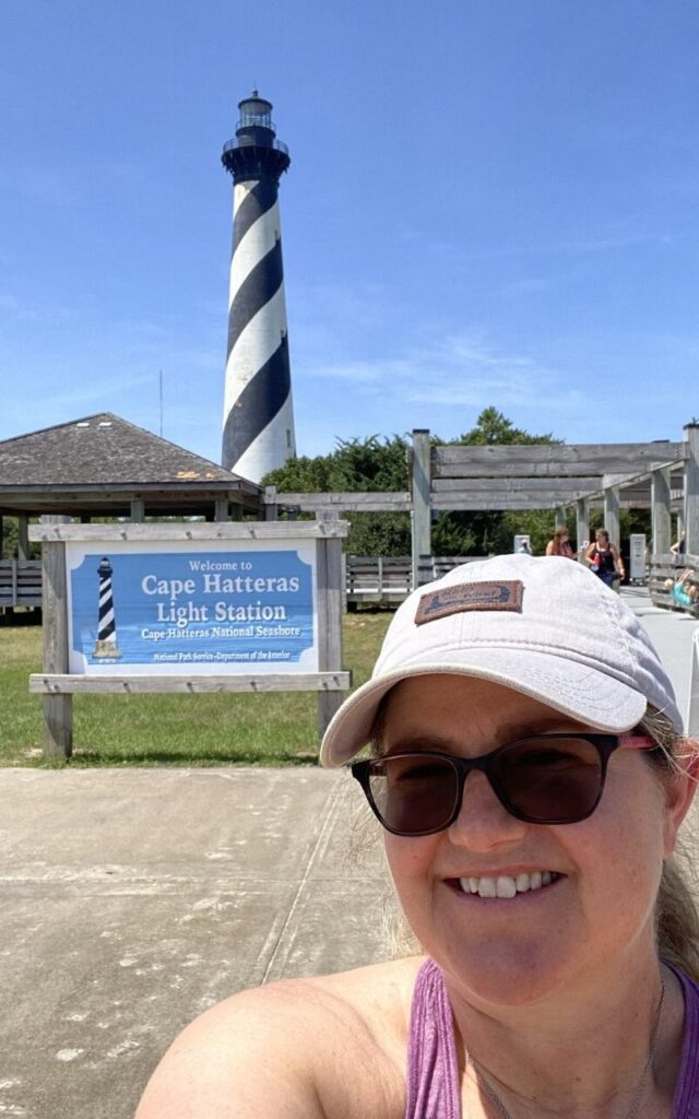 Shannon Winakur takes a selfie in front of the sign for the Cape Hatteras Light Station. The black and white diagonally striped Cape Hatteras Lighthouse can be seen in the background. 