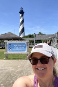 Shannon Winakur takes a selfie in front of the sign for the Cape Hatteras Light Station. The black and white diagonally striped Cape Hatteras Lighthouse can be seen in the background.